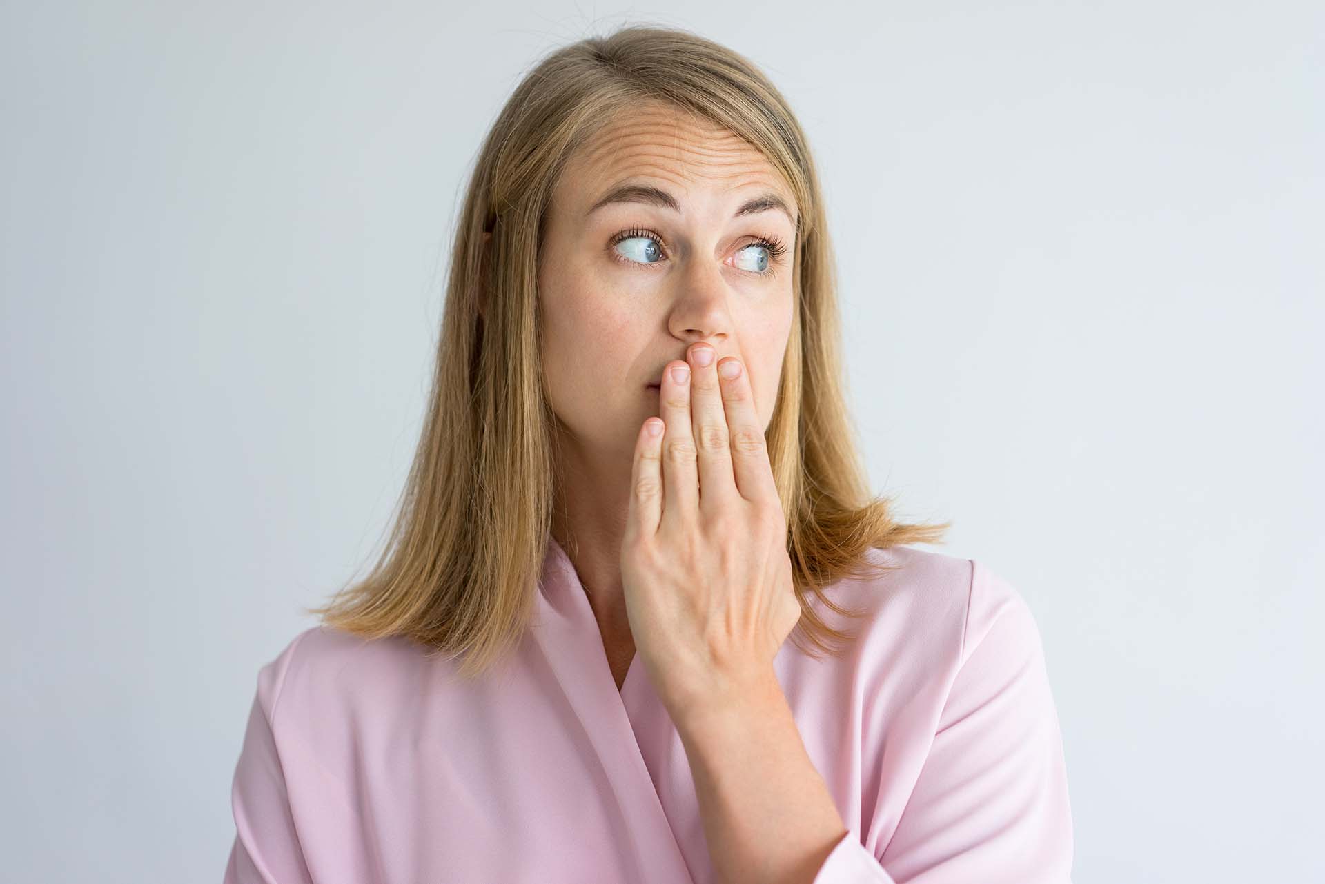 Portrait of shocked young Caucasian woman wearing pink blouse covering mouth with hand. Fear, bad breath concept