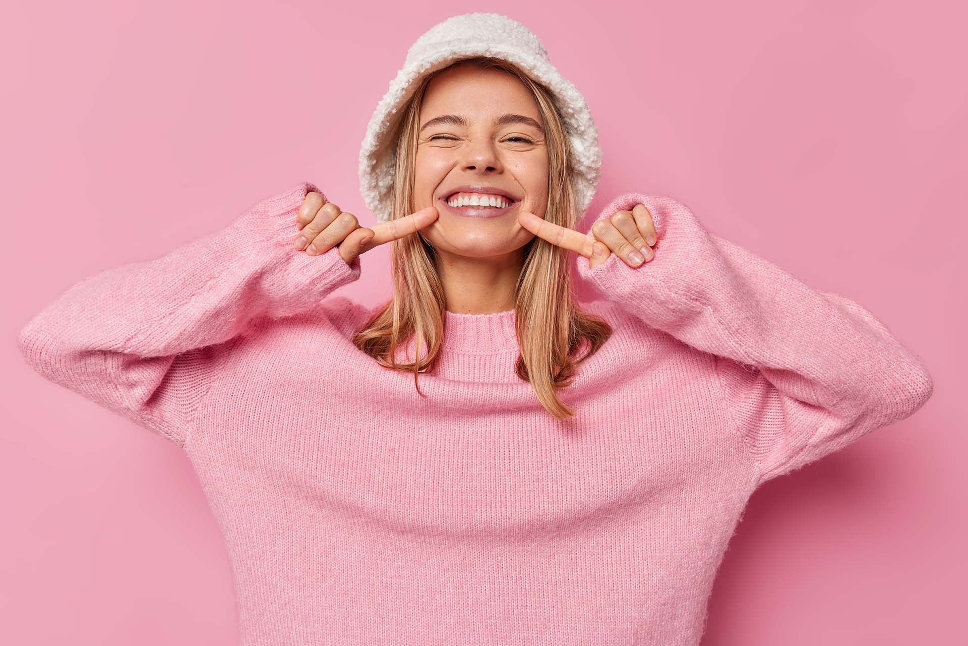 Positive fair haired woman points at her white teeth smiles broadly wears fashionable panama and casual jumper isolated over pink background has happy mood