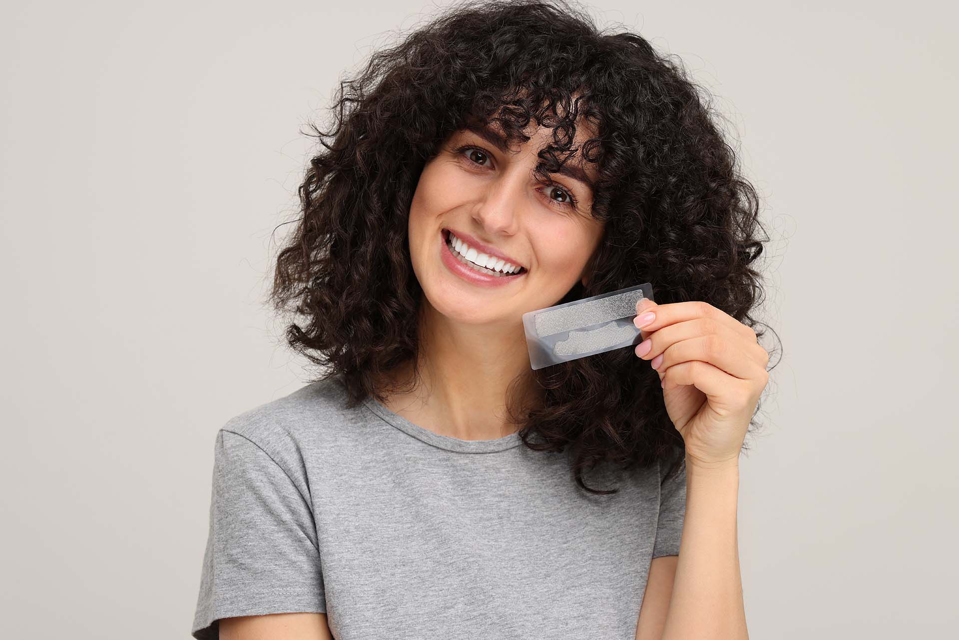 Young woman holding teeth whitening strips on light grey background