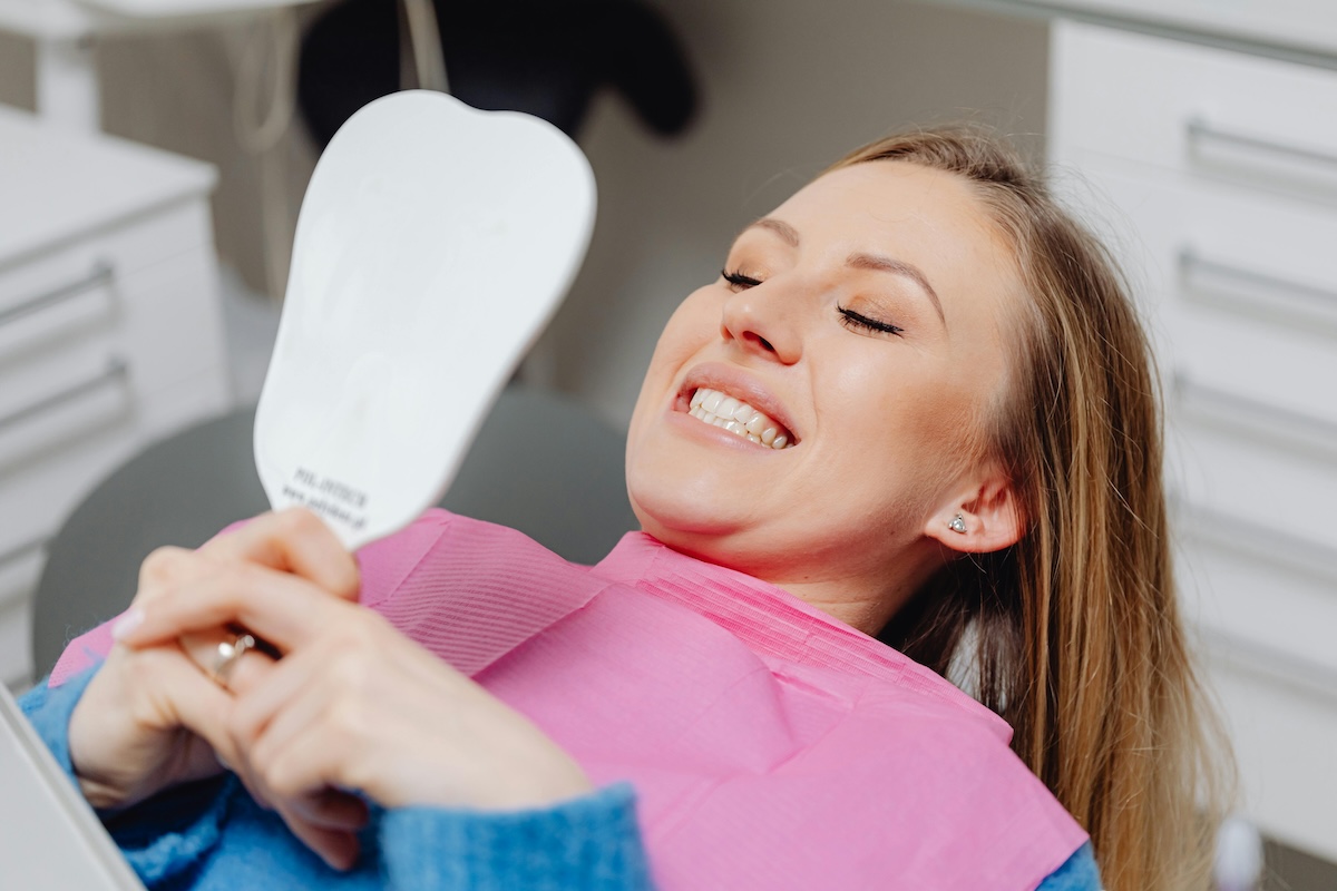 A happy female patient smiling while looking at her teeth whitening results in a hand mirror at a dental office.