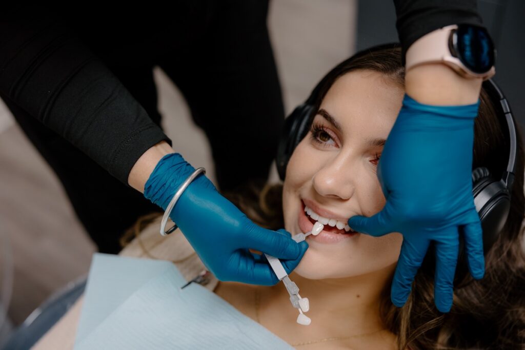 A dental professional in blue gloves holds a tooth shade guide next to a smiling female patient during a teeth whitening consultation at a dental office.
