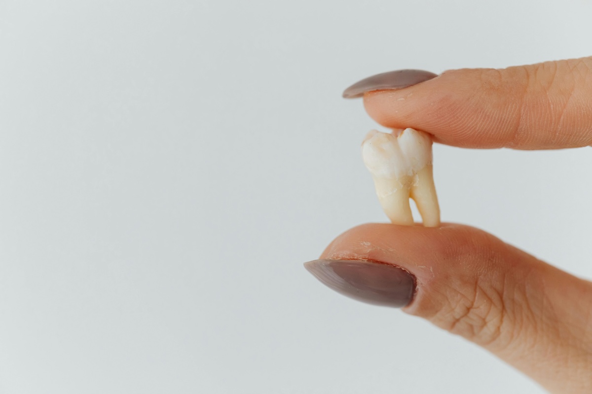 A hand holding a knocked-out tooth by the root against a light gray background.