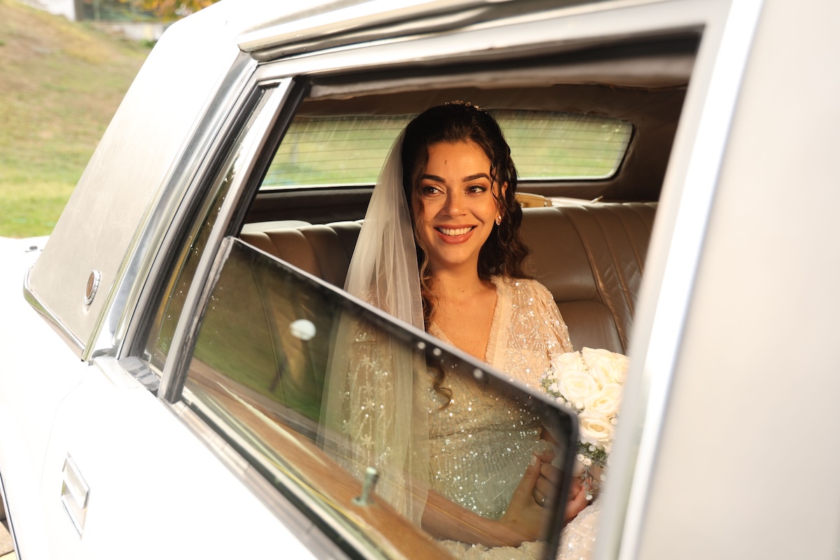 Bride smiling with visible teeth in a vintage wedding car, wearing a veil and holding a bouquet