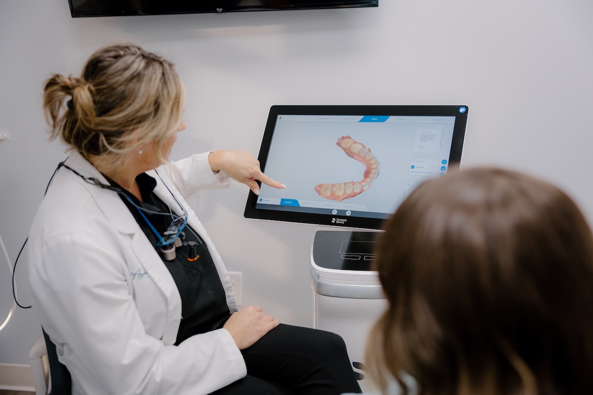 Dentist in a white coat pointing to a 3D digital scan of a patient's teeth on a monitor while explaining it to the patient