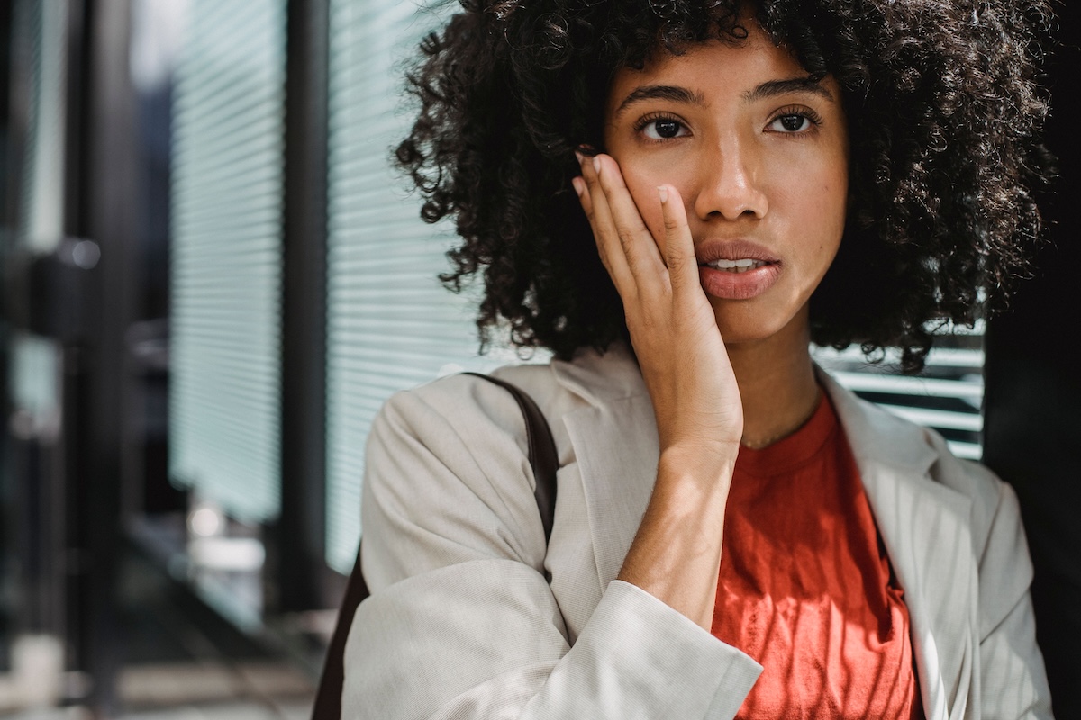 Woman with curly hair holding her hand to her cheek and jaw with a pained expression while standing in an office