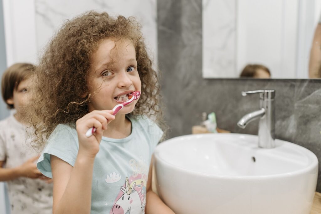 Smiling girl with curly hair brushing her teeth at a bathroom sink as part of a daily routine for healthy gums