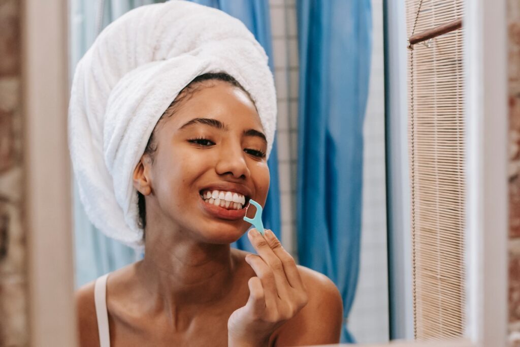 Smiling woman with a towel on her head using a floss pick on her lower teeth in front of a bathroom mirror