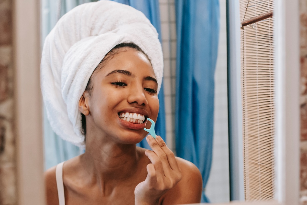 Smiling woman with a towel on her head using a floss pick on her lower teeth in front of a bathroom mirror