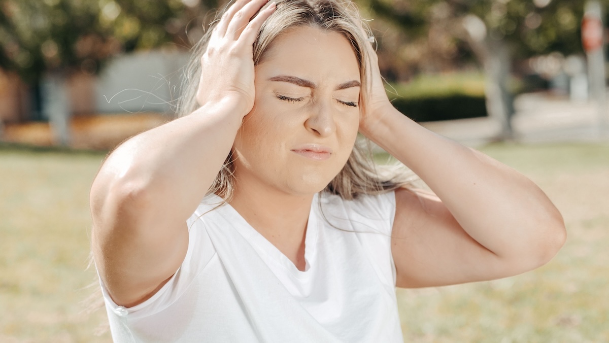 Woman grimacing with both hands pressed to the sides of her head, a sign of headache or ear pressure that can come with TMJ