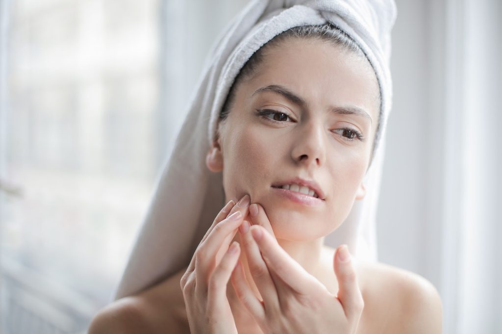 Woman with a towel wrapped around her hair pressing her fingertips against her cheek near her mouth, looking uncomfortable, as if feeling for a swollen gum around one tooth.
